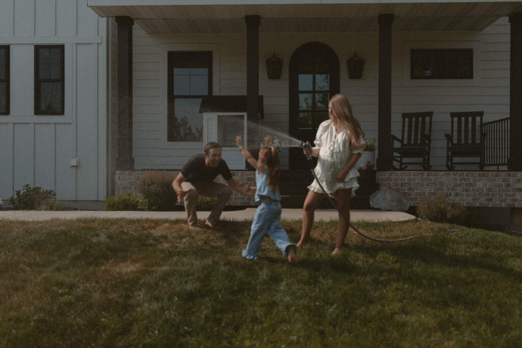 Mom spraying a hose while toddler runs through it at outdoor family photo session in Minnesota
